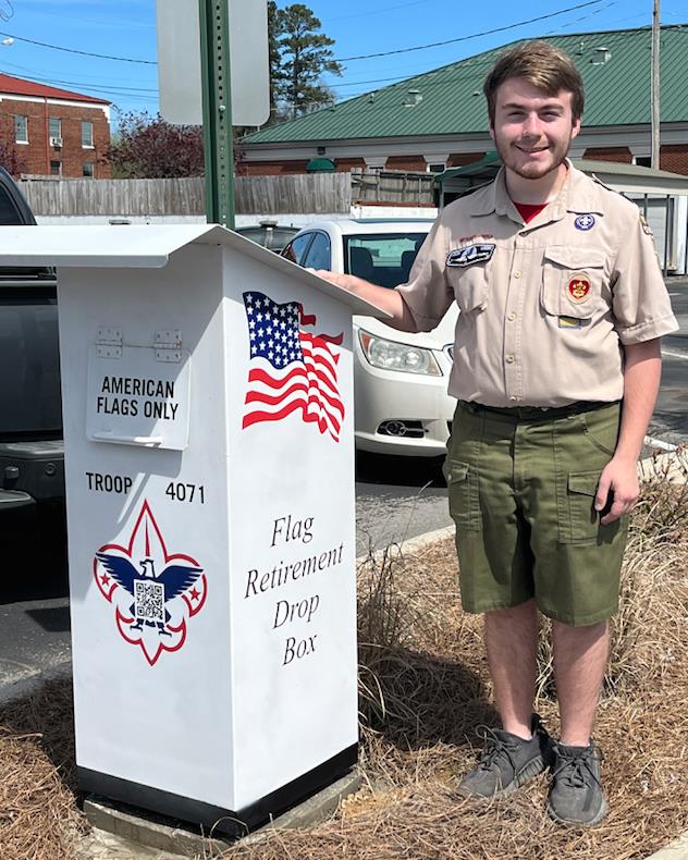 Young man standing in his boy scout uniform next to a a flag retirement drop box.